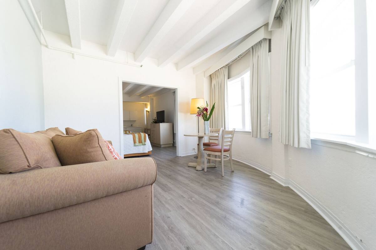 A modern hotel room with a beige sofa, a dining table with chairs, a lamp, and a view of the hallway.