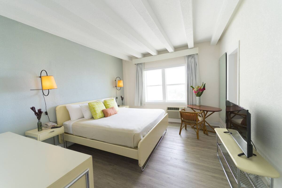 A hotel room with a bed, bedside lamps, a desk, a chair, a table, a TV, and a window with curtains.