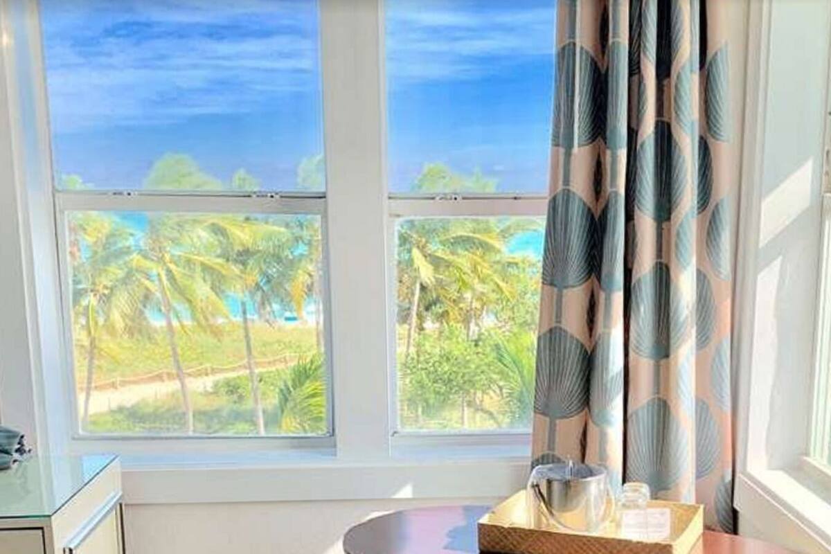 A room with a view of palm trees and a beach, a window with curtains, and a tray with a metal pitcher and glasses.