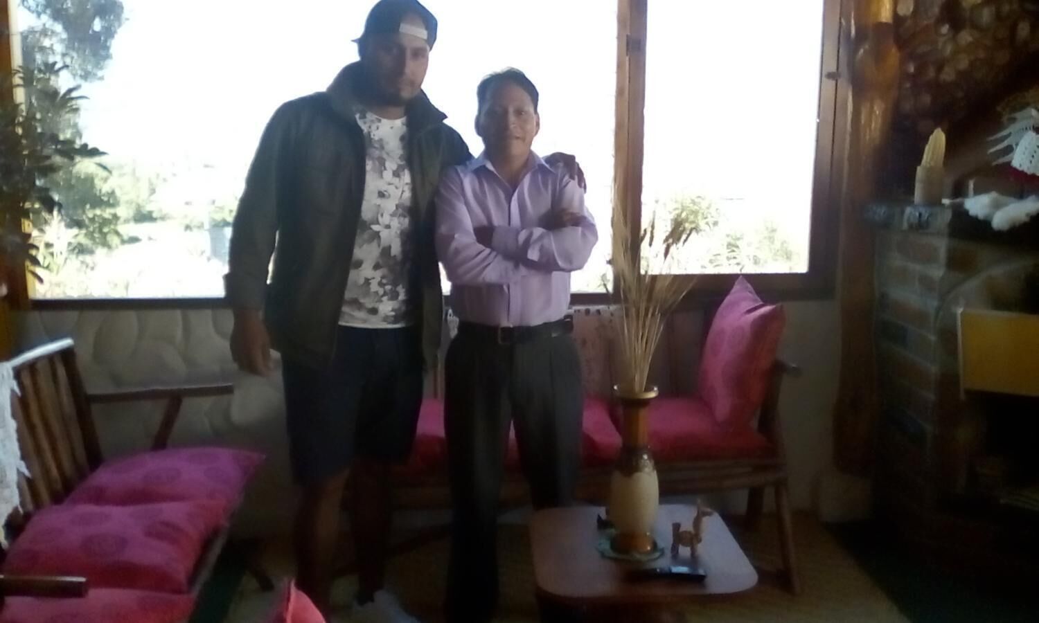 Two people standing in a room with a large window, a pink chair, and a small table with a vase.