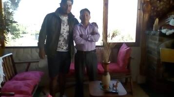 Two people standing in a room with a large window, a pink chair, and a small table with a vase.