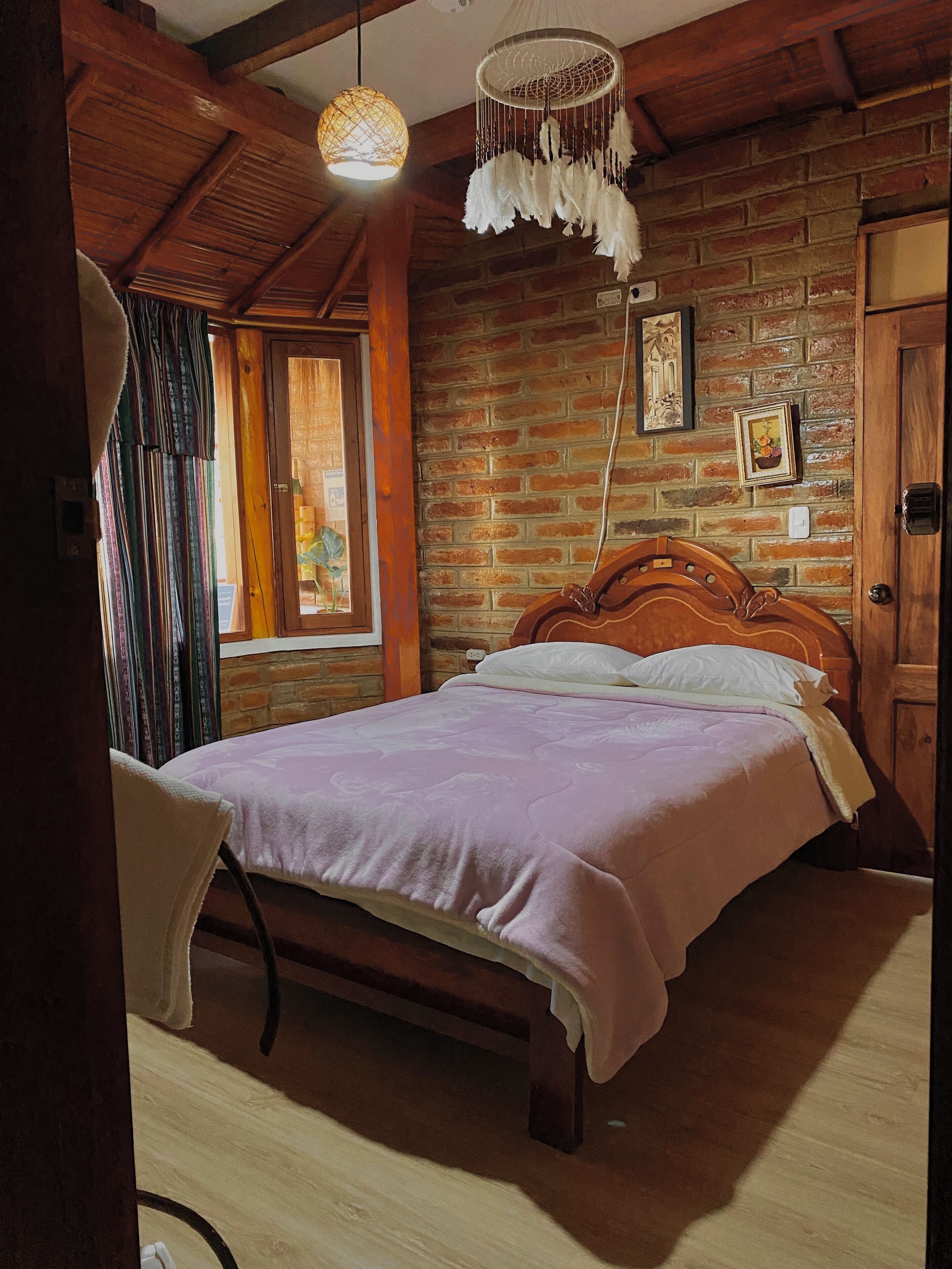 Una habitación con estructura de madera, una cama, una silla, una lámpara colgante y una pared de ladrillo.