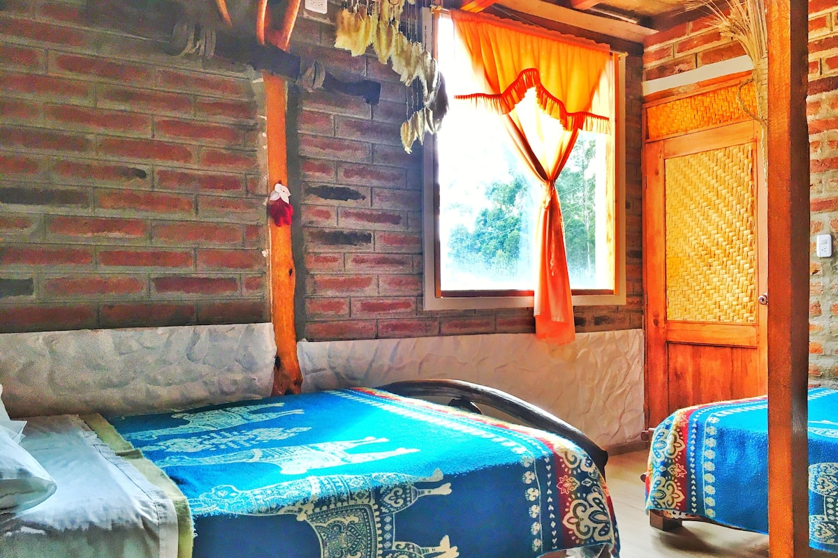 A rustic room with a brick wall, a wooden door, and a window with orange curtains. There are two beds with colorful bedspreads.
