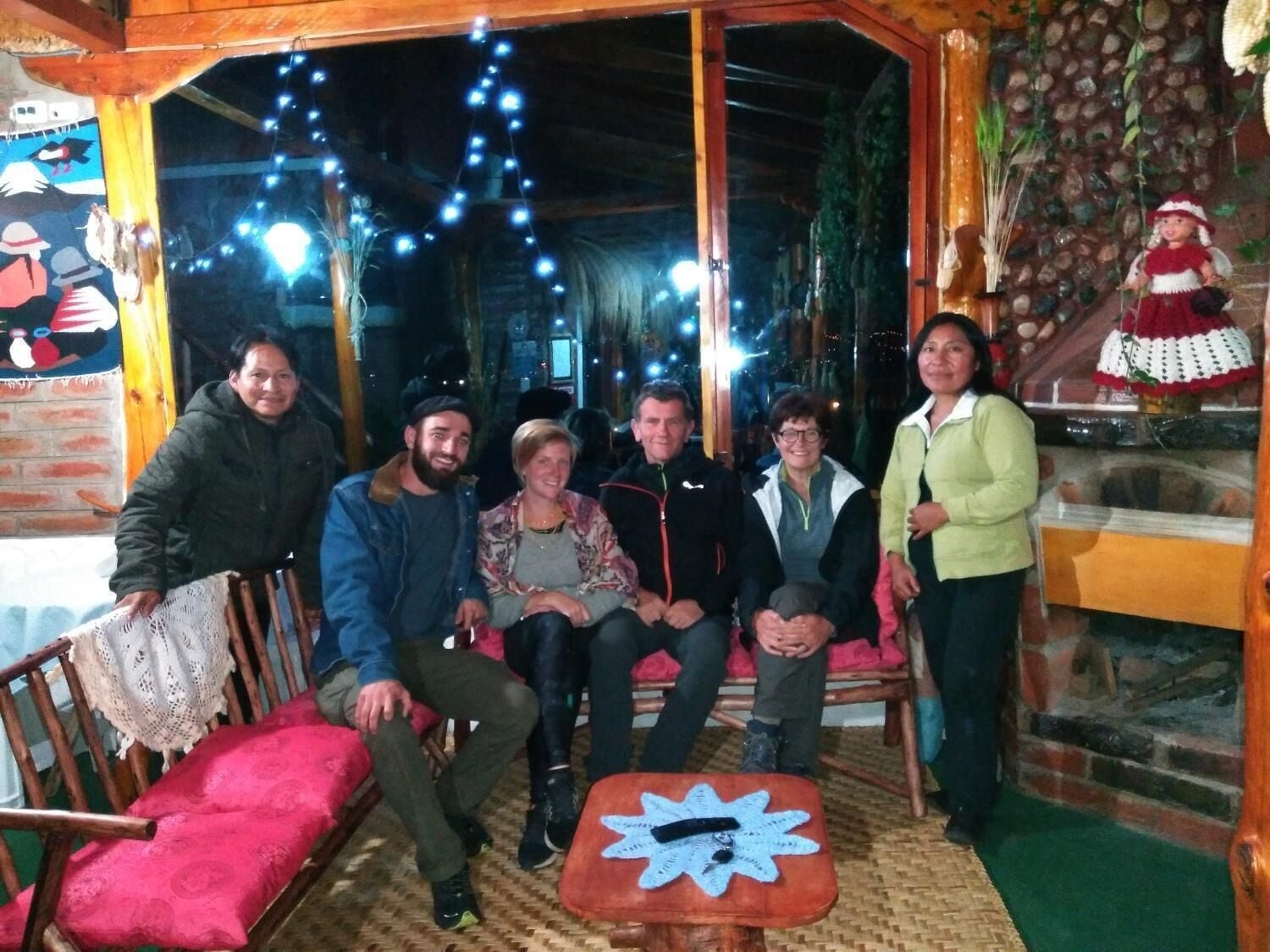 Un grupo de personas sentadas y de pie en una habitación acogedora, con luz cálida, muebles de madera y luces de estilo guirnalda.