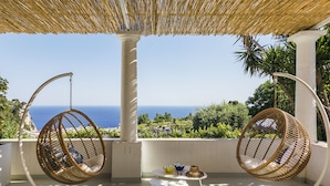 A balcony with two wicker hanging chairs, a small round table, and a view of the sea. - Suite Time Capri Villa La Pergola (Capri)