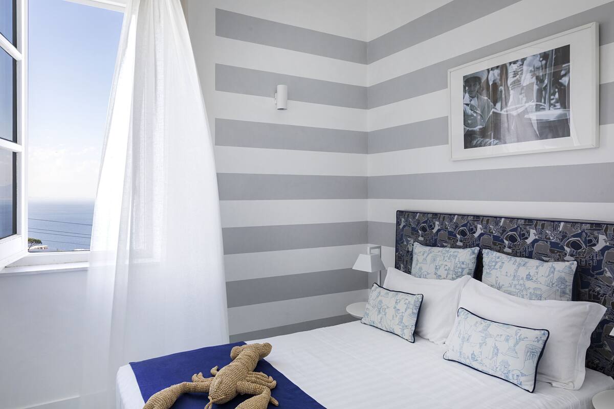 A hotel room with a striped wall, a bed with blue and white pillows, a window with a view, and a framed picture on the wall.