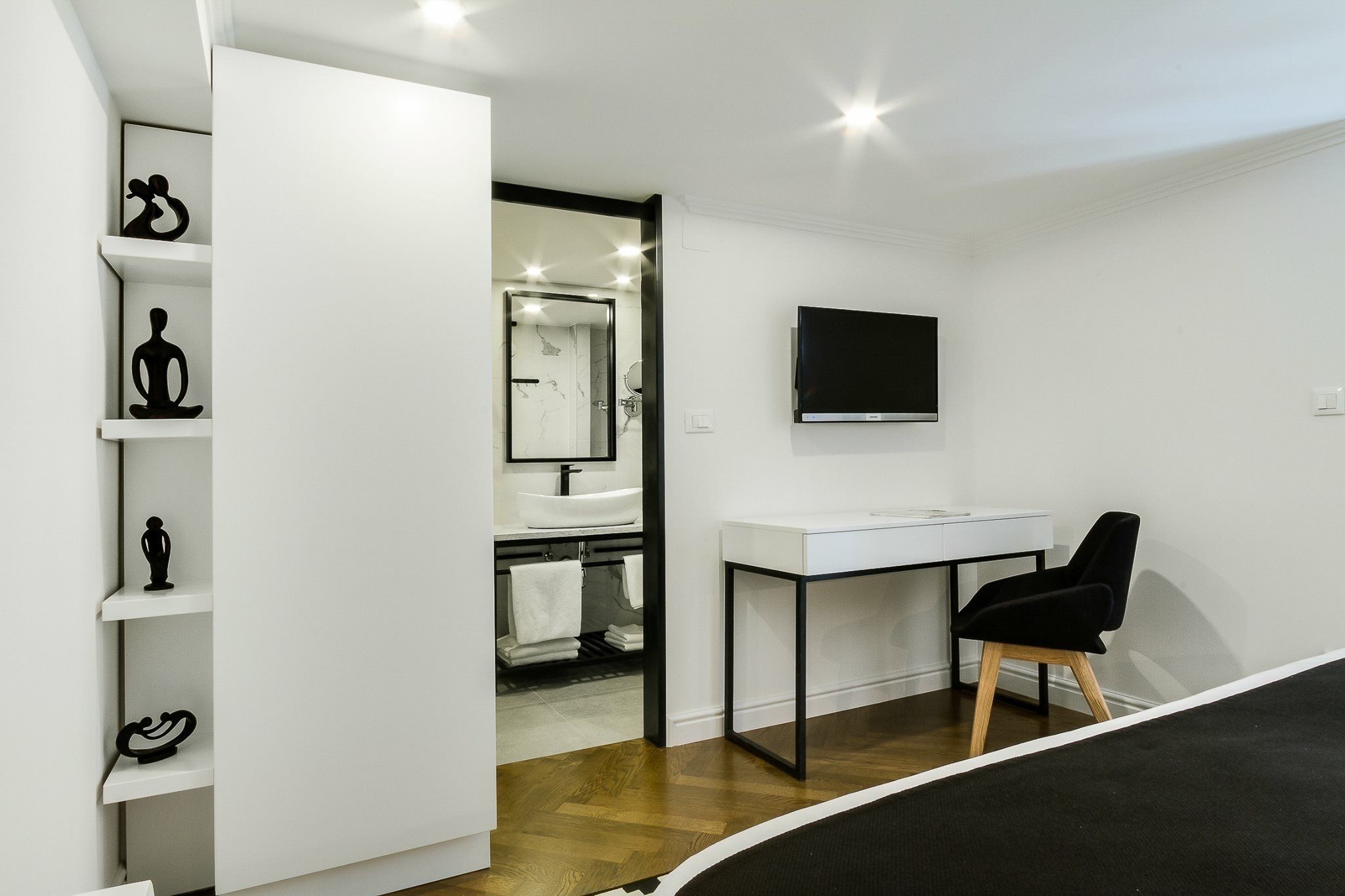 A modern hotel room with a white desk, black chair, and a TV mounted on the wall.