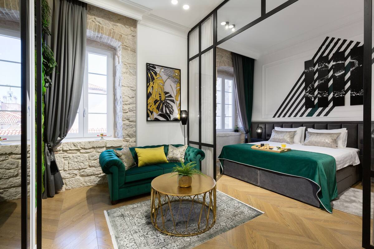 Ein modernes Schlafzimmer mit einem grünen Sofa, einem runden goldenen Tisch und einem Bett mit grüner Bettdecke.