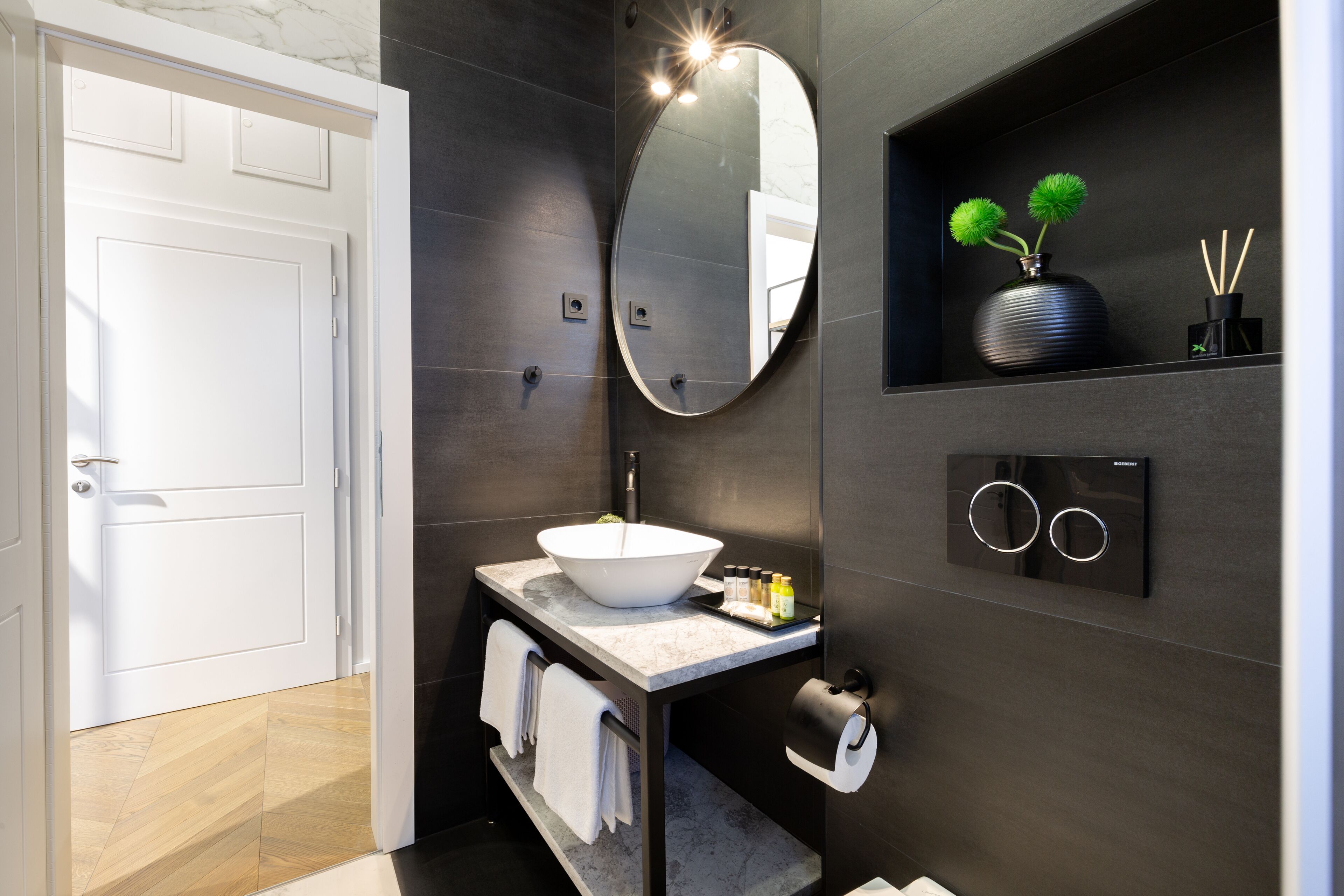 Un baño moderno con un lavabo blanco, baldosas oscuras, un espejo grande y un estante con objetos decorativos.