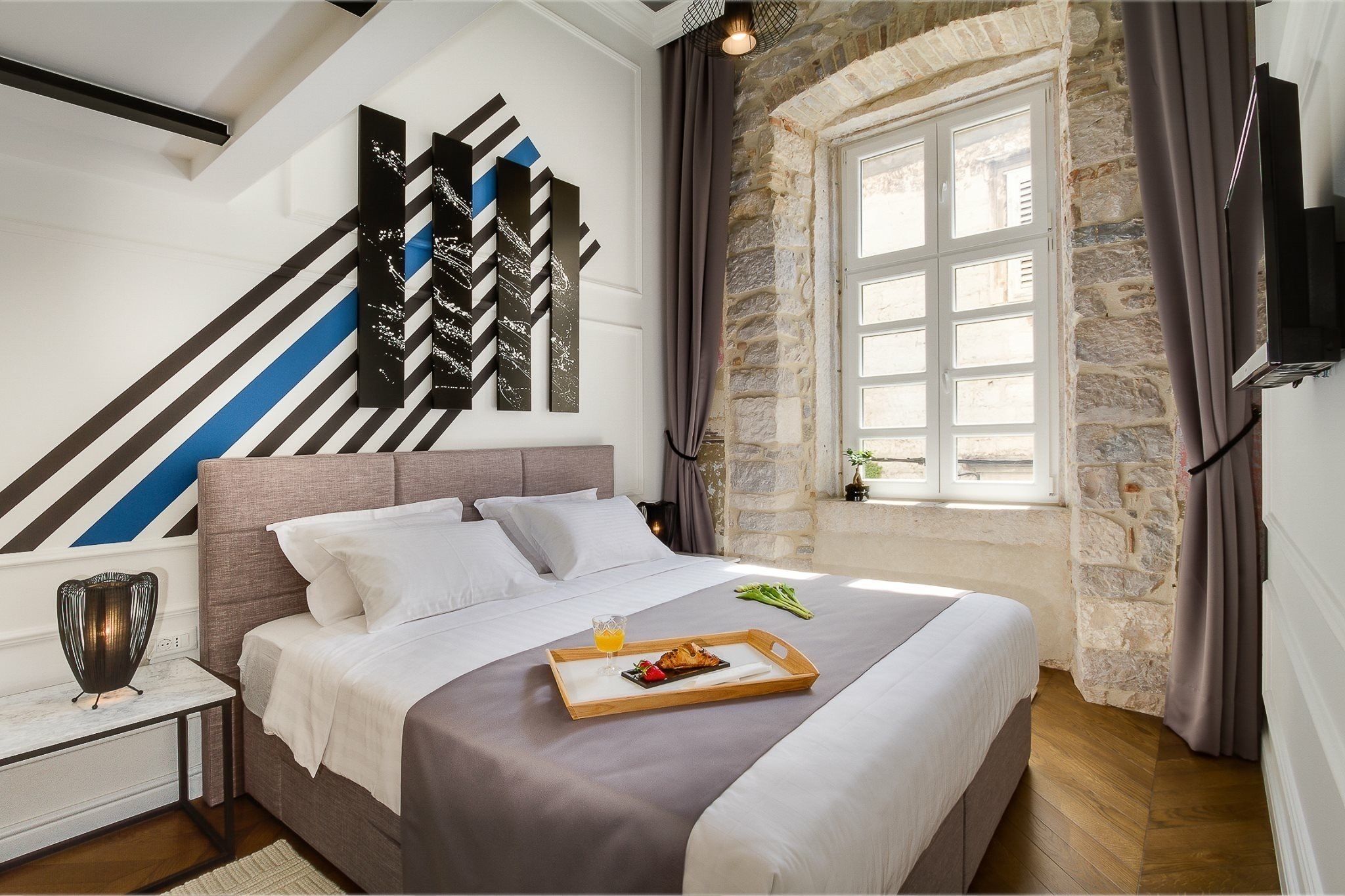 A neatly made bed with a tray of breakfast, a stone wall, and a large window.