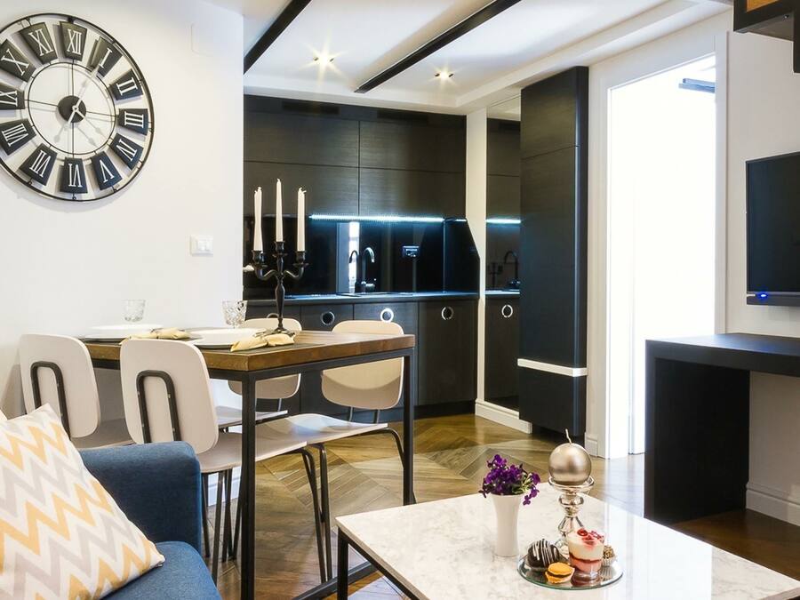 A modern living room with a large wall clock, a dining area with a table and chairs, and a kitchen with dark cabinetry and a TV.
