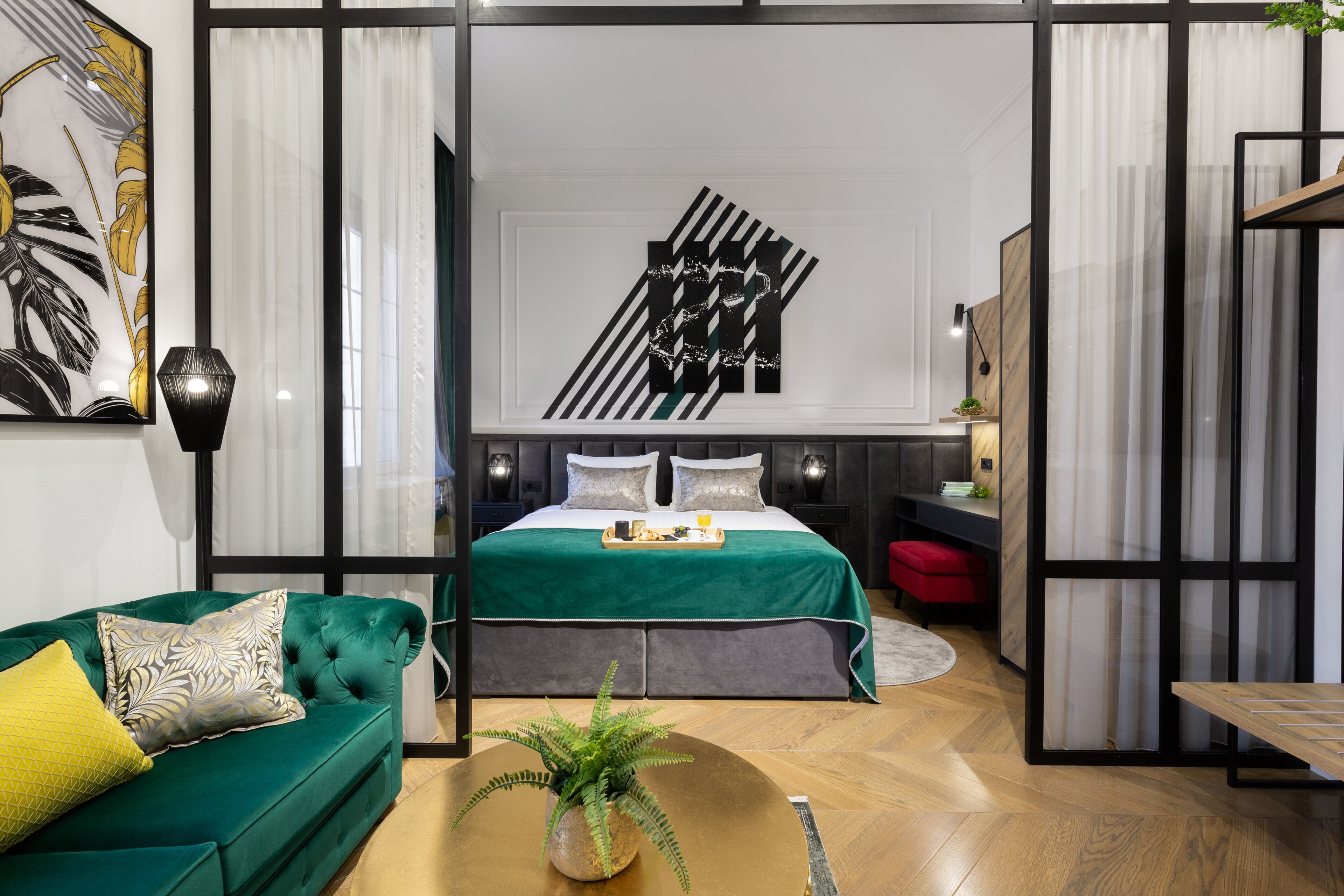 Un dormitorio moderno con un sofá verde, una cama con edredón verde, una obra de arte en la pared y una pequeña mesa con una planta.
