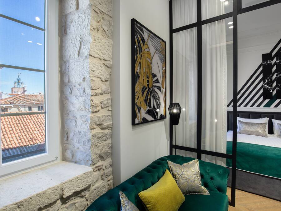Una habitación con pared de piedra, ventana con vistas a edificios, sofá verde con cojines y cama con colcha verde.