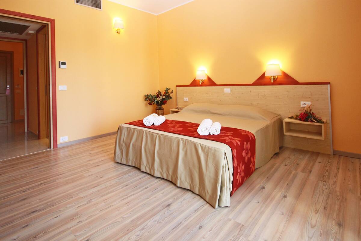 A hotel room with a wooden floor, a bed with a red and beige bedspread, two bedside lamps, and a nightstand with a vase of flowers.