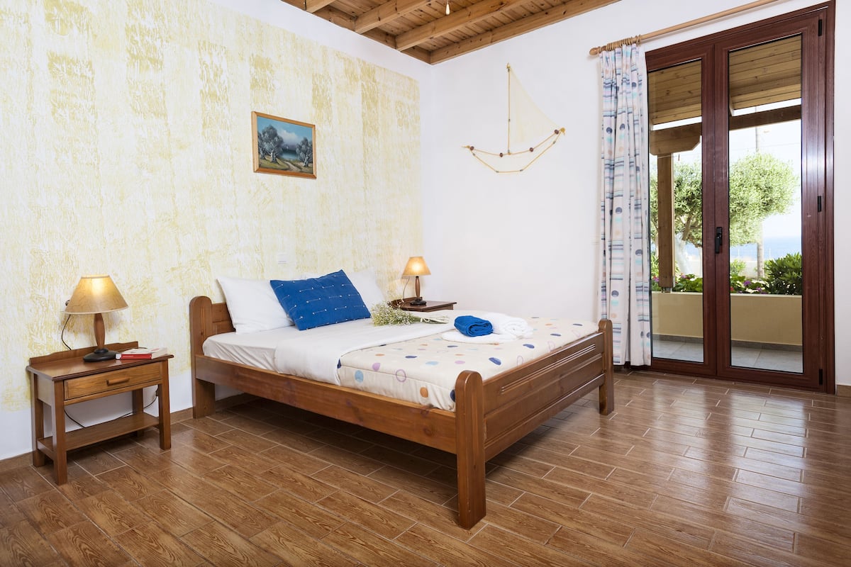 Une chambre avec un lit en bois, deux tables de chevet avec des lampes, un tableau au mur et une vue à travers une porte vitrée.