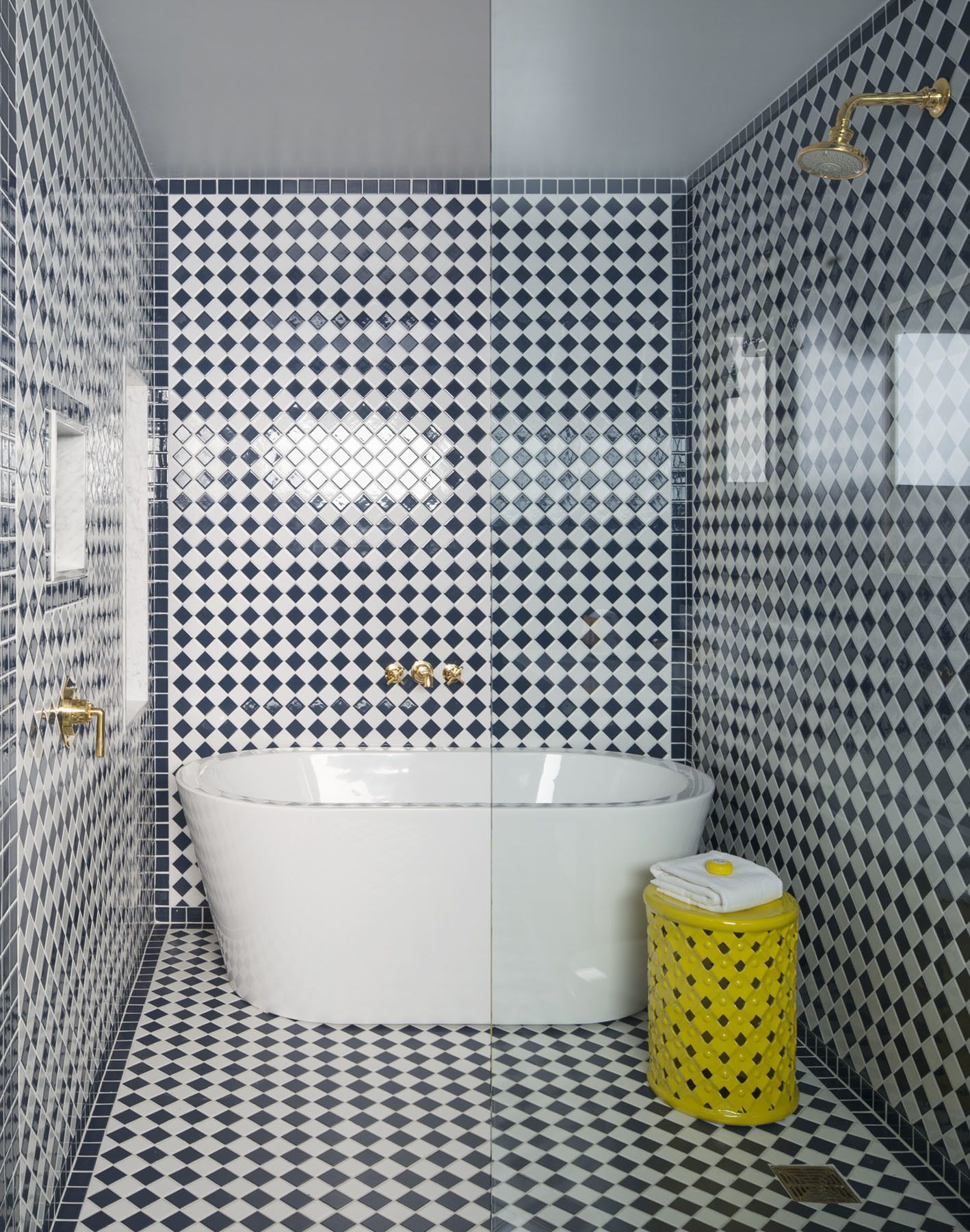 A bathroom with a large bathtub, a yellow stool, and diamond-patterned tiles on the walls and floor.