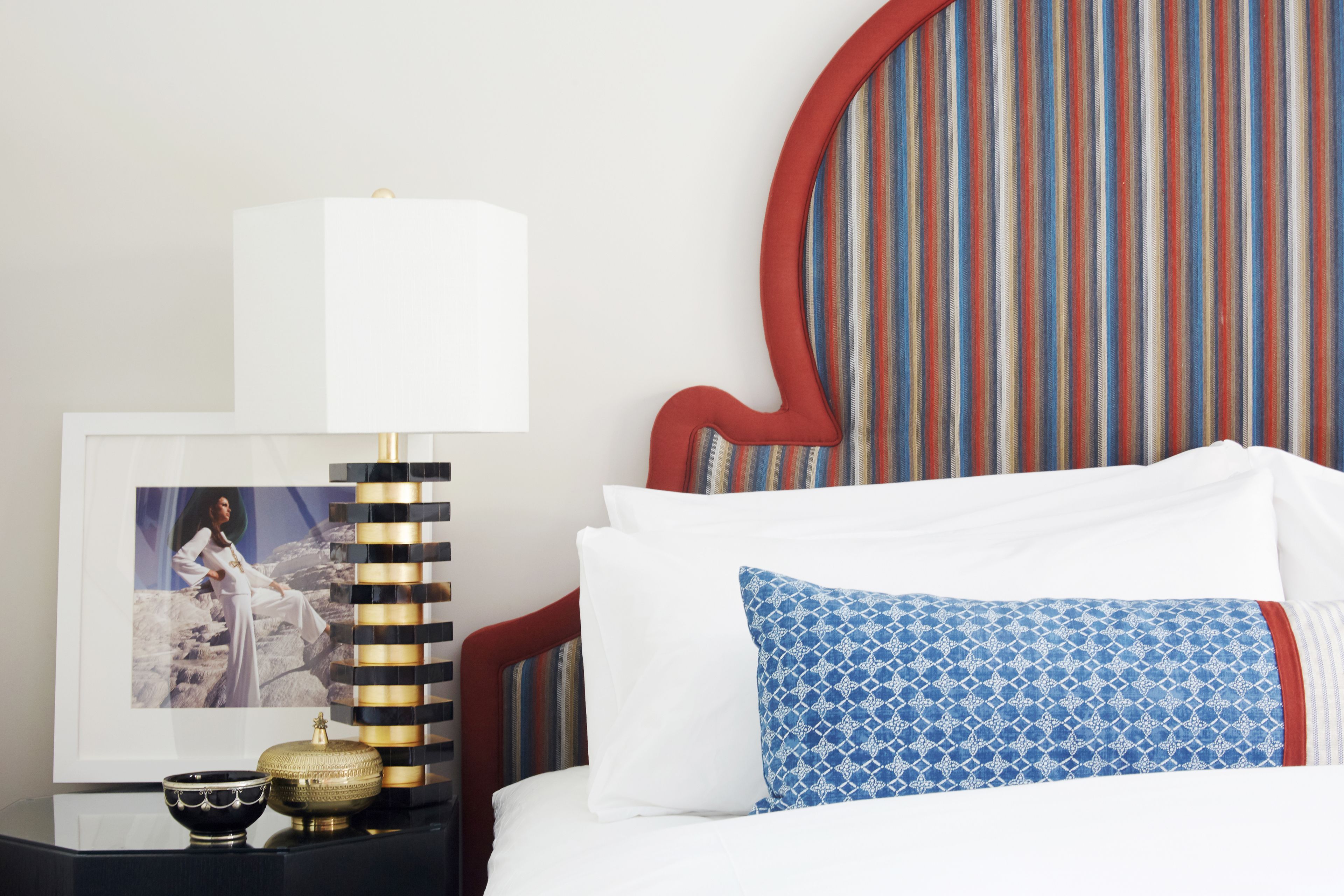 A neatly made bed with white and blue patterned pillows, a framed picture on a bedside table, and a striped wall behind.