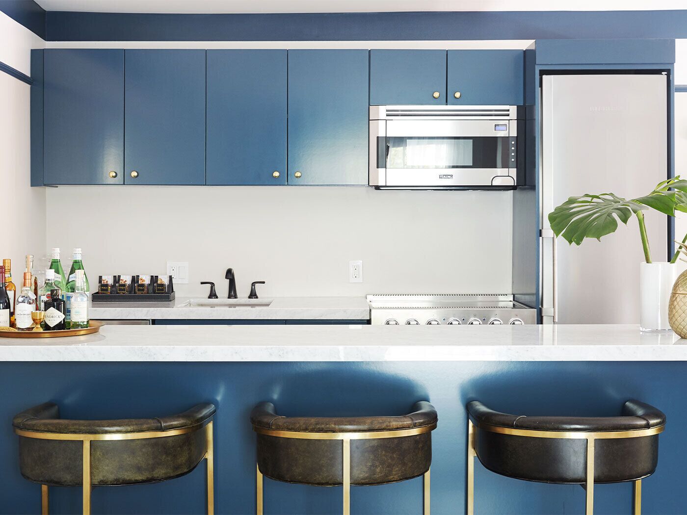Una cucina moderna con pensili blu, un forno a microonde in acciaio inossidabile e un piano di lavoro bianco. Sono presenti tre sgabelli da bar con dettagli dorati e un vaso con una pianta sul bancone.