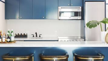 A modern kitchen with blue cabinets, a stainless steel microwave, and a white countertop. There are three bar stools with gold accents and a potted plant on the counter.