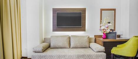 Habitación de hotel con sofá color beige, televisor de pantalla plana montado en la pared, escritorio con una revista, un florero con flores y una silla amarilla.