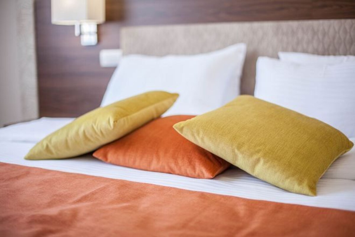 Una cama con ropa de cama blanca y naranja, dos almohadas y una lámpara fijada a la pared.
