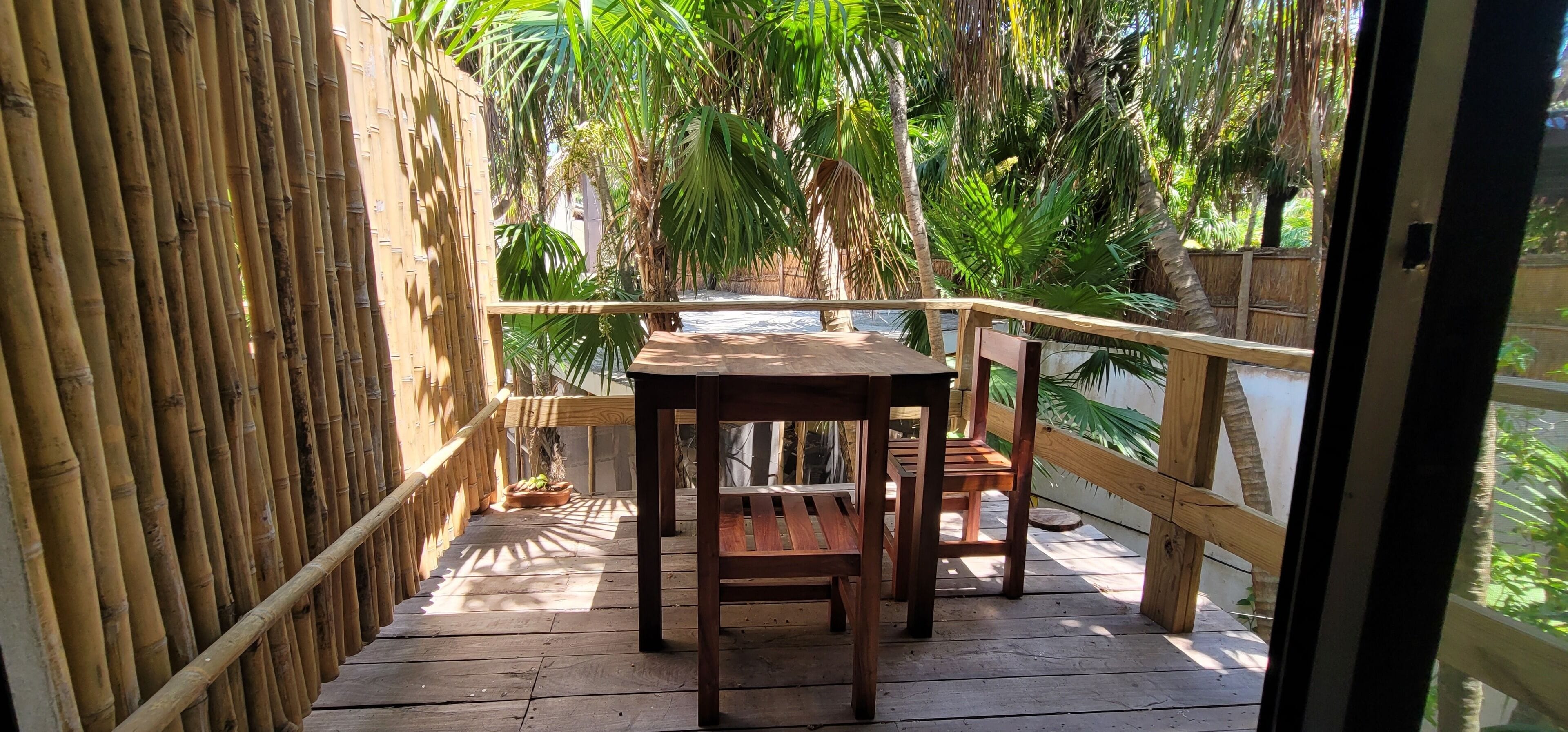 A wooden deck with a table and chairs, surrounded by bamboo fencing and lush tropical plants.