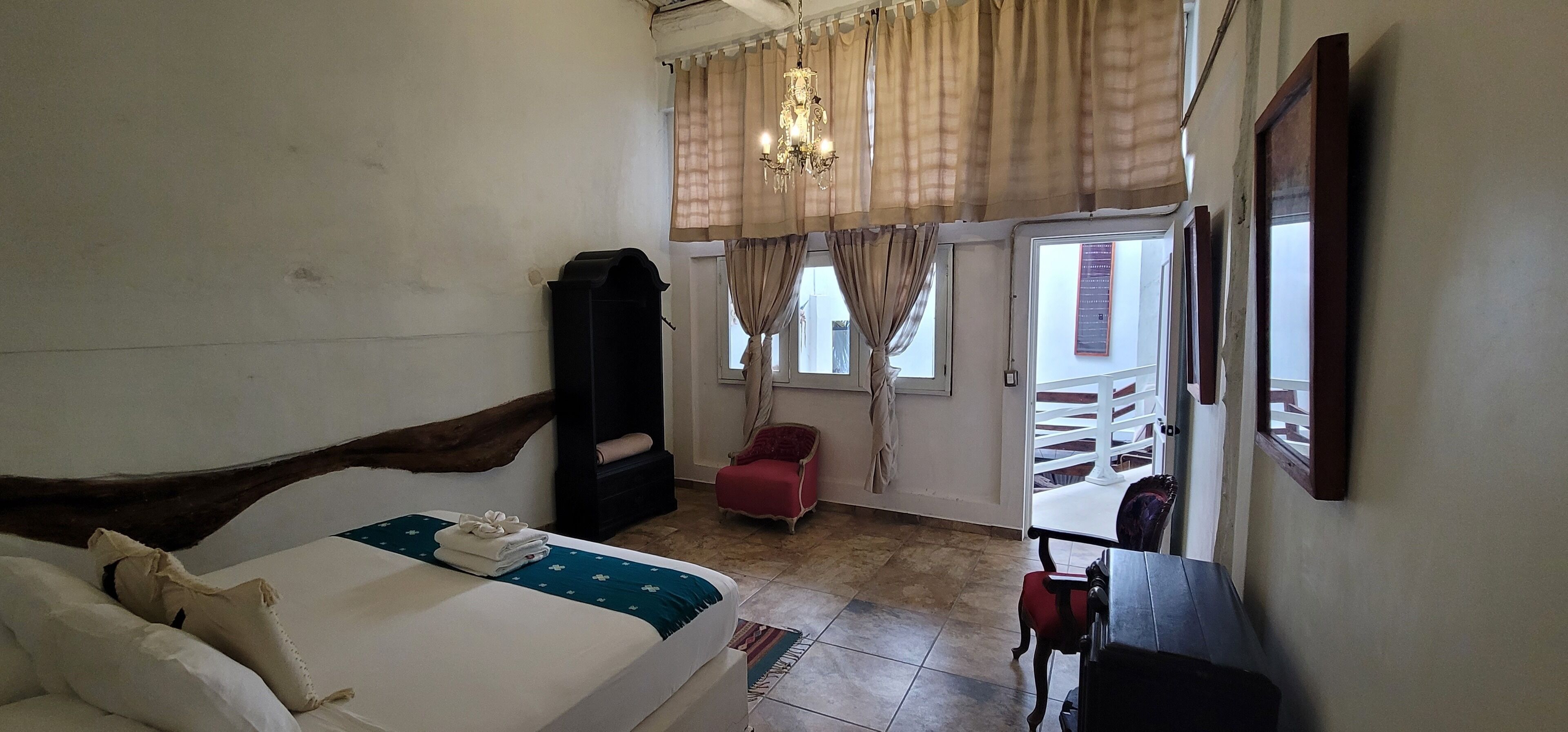 Un dormitorio con cama, armario, una silla roja, una mesa y una ventana con cortinas.