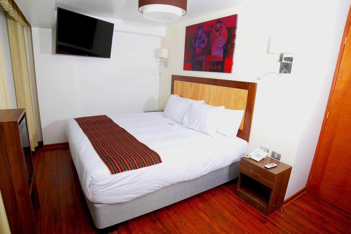 A hotel room with a bed, a TV mounted on the wall, a painting, a bedside table with a phone, and a wooden floor.