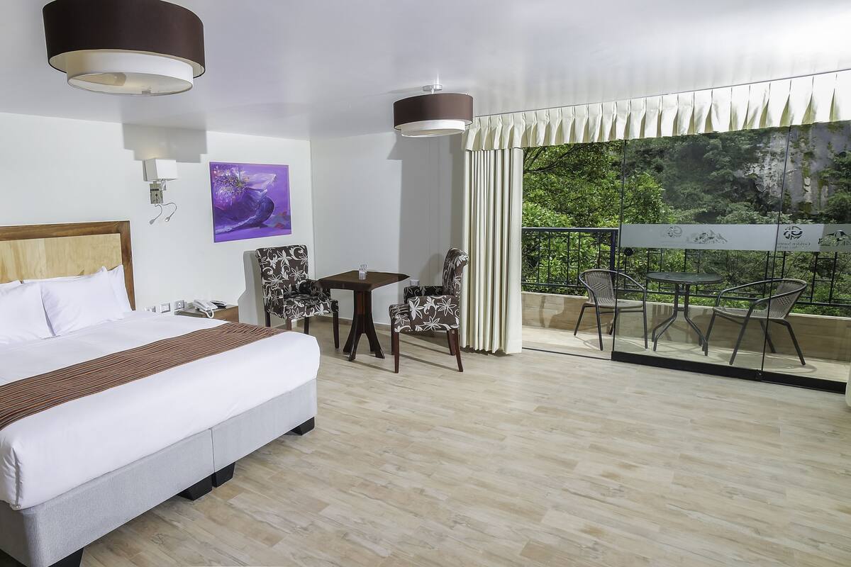 A hotel room with a large bed, a small table with chairs, a balcony with a view of greenery, and a painting on the wall.