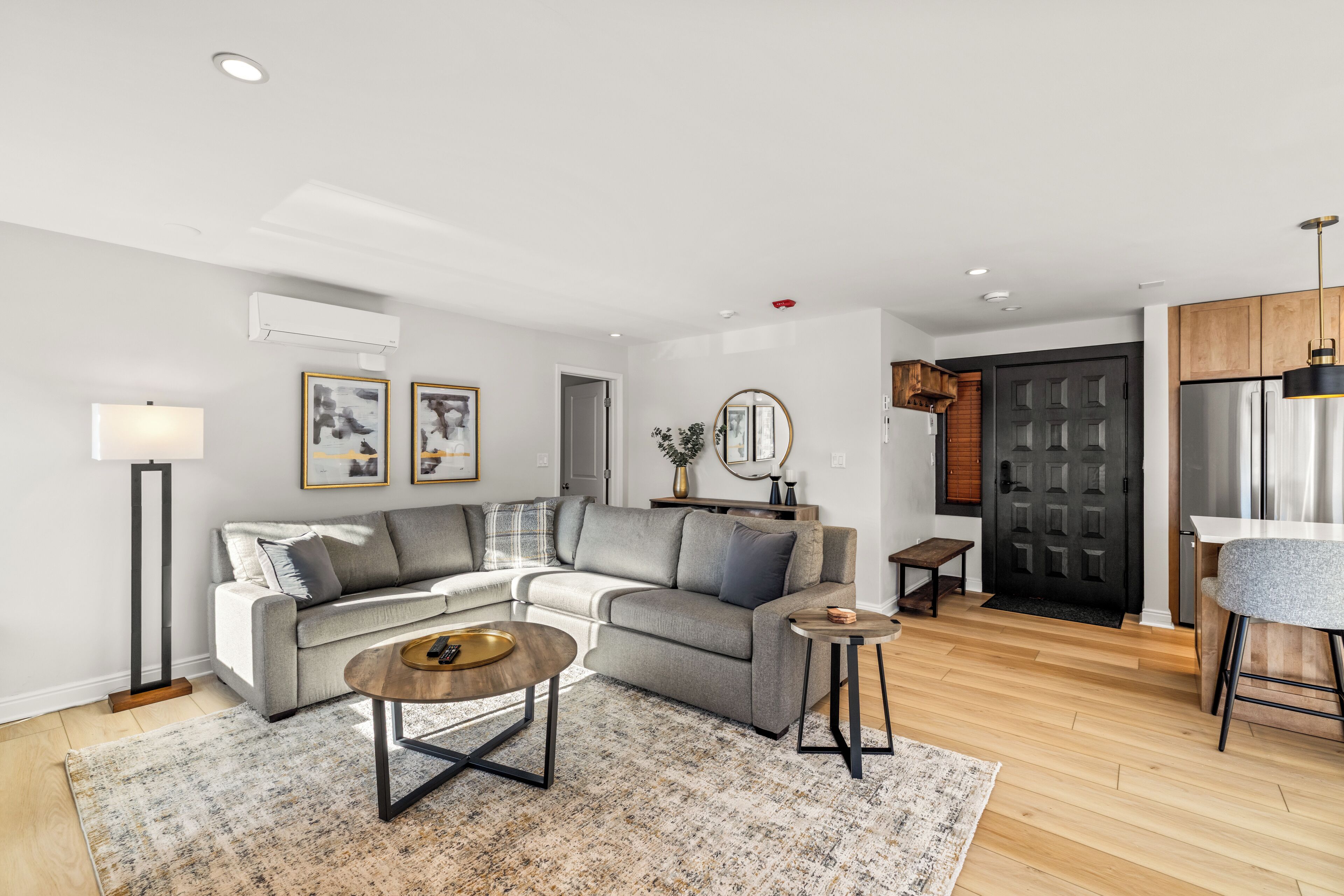 A modern living room with a grey sectional sofa, a round coffee table, and a small side table. There is a floor lamp, a mirror, and framed artwork on the wall.