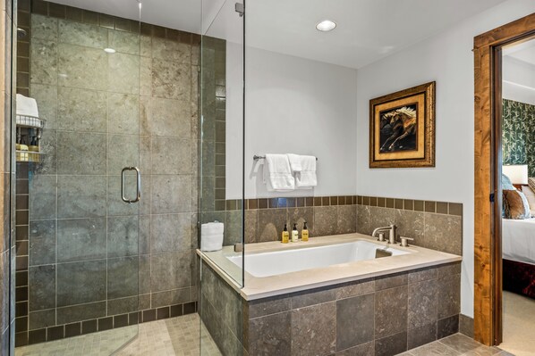 A modern bathroom with a glass-enclosed shower, a large bathtub, and a wooden door leading to a bedroom.