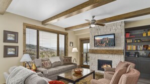 A living room with a stone fireplace, a television mounted above it, and a bookshelf filled with books and decorative items. - Edgemont 2402 Luxury On Mountain Condo With Ski-In/Ski-Out Access! (Steamboat Springs)