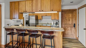 Una cocina con armarios de madera, un refrigerador de acero inoxidable, una encimera de mármol y tres taburetes de bar.