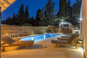 A swimming pool with lounge chairs, a wooden deck, and a covered area with a barbecue.