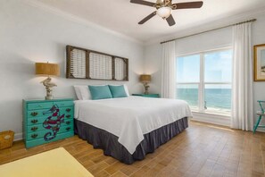 A bedroom with a bed, two nightstands, a ceiling fan, a window with a view of the ocean, and a decorative wall piece.
