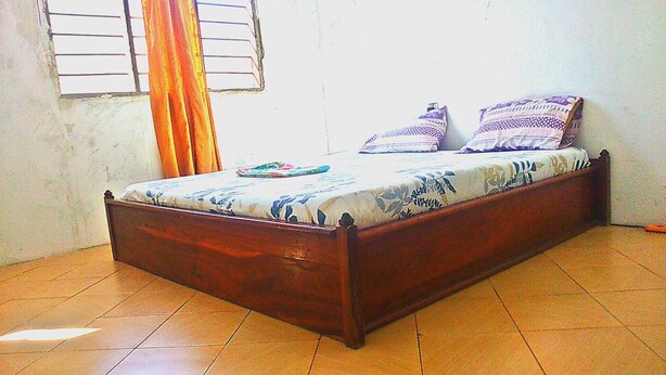 A single bed with a floral bedspread, a wooden headboard, and a window with blinds and a curtain. - Homestay in Dar es salaam (DSM)