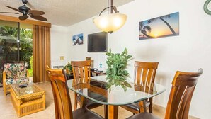 A dining area with a glass table surrounded by wooden chairs, a ceiling fan, and a wall-mounted TV. - Open Design Suite w/Modern Kitchen, Lanai to Lawn, WiFi–Kamaole Sands 9106 (KIHEI)
