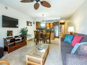A living room with a television, a dining area with chairs and a table, a kitchen with cabinets and appliances, and a sofa with cushions.