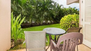 A patio with a round table and two chairs, surrounded by lush greenery and palm trees. - Open Design Suite w/Modern Kitchen, Lanai to Lawn, WiFi–Kamaole Sands 9106 (KIHEI)