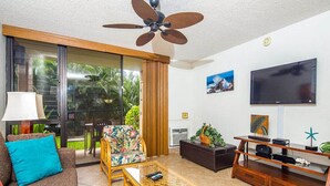 A living room with a sofa, a wicker coffee table, a television, and a ceiling fan. - Open Design Suite w/Modern Kitchen, Lanai to Lawn, WiFi–Kamaole Sands 9106 (KIHEI)