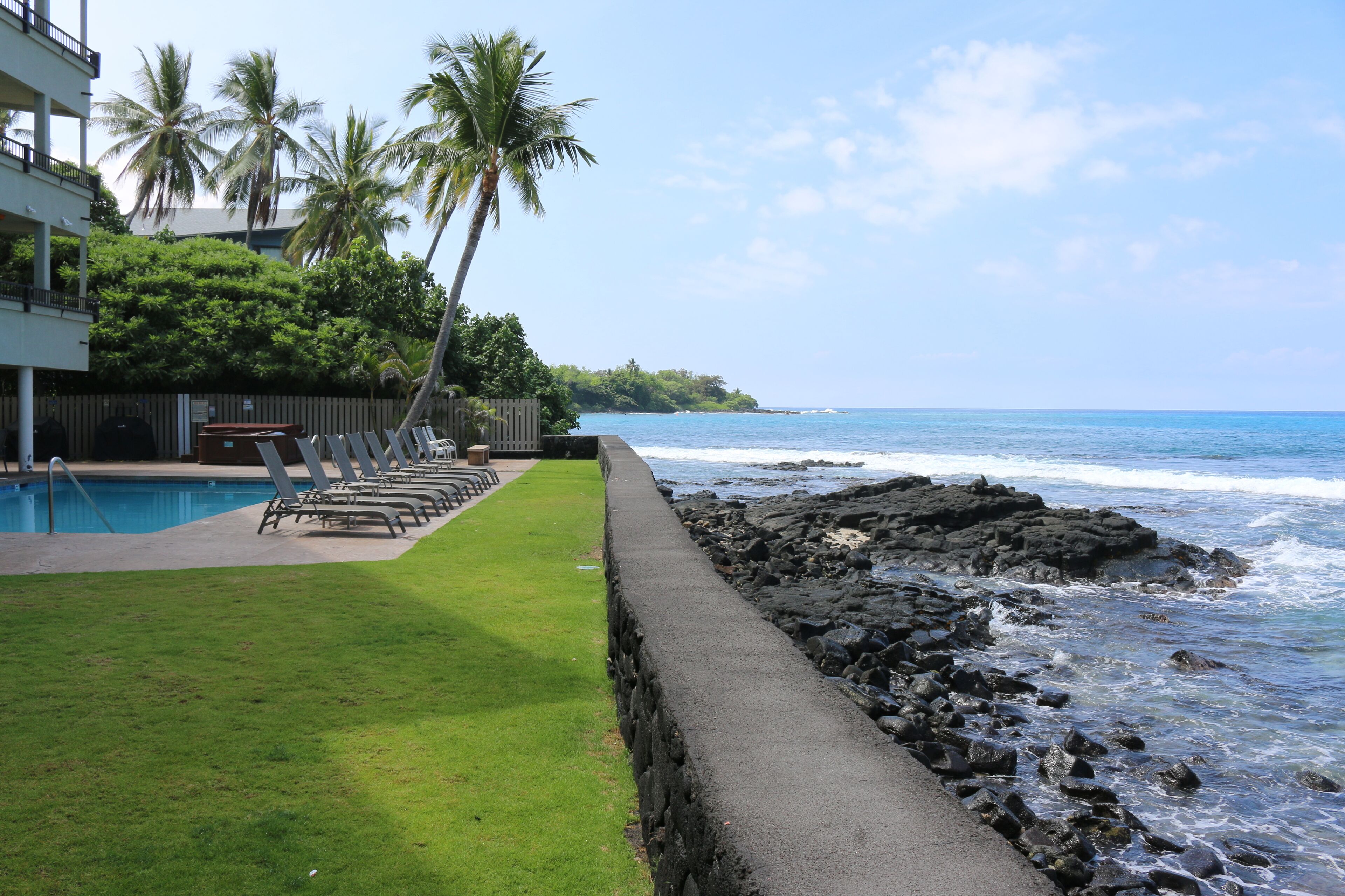 A pool area with lounge chairs, a concrete pathway, and a rocky shoreline with waves.