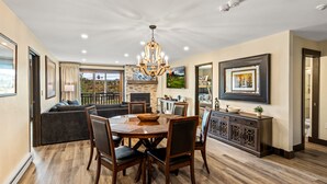 A dining area with a chandelier, wooden table, and chairs. There is a sofa and a cabinet with decorative items.