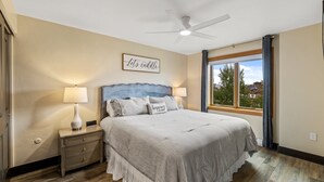 A bedroom with a bed, nightstands, a ceiling fan, a window with curtains, and a view of the outside.