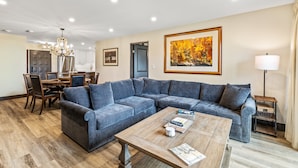 A modern living room with a sectional sofa, a wooden coffee table, and a dining area with chairs and a chandelier.