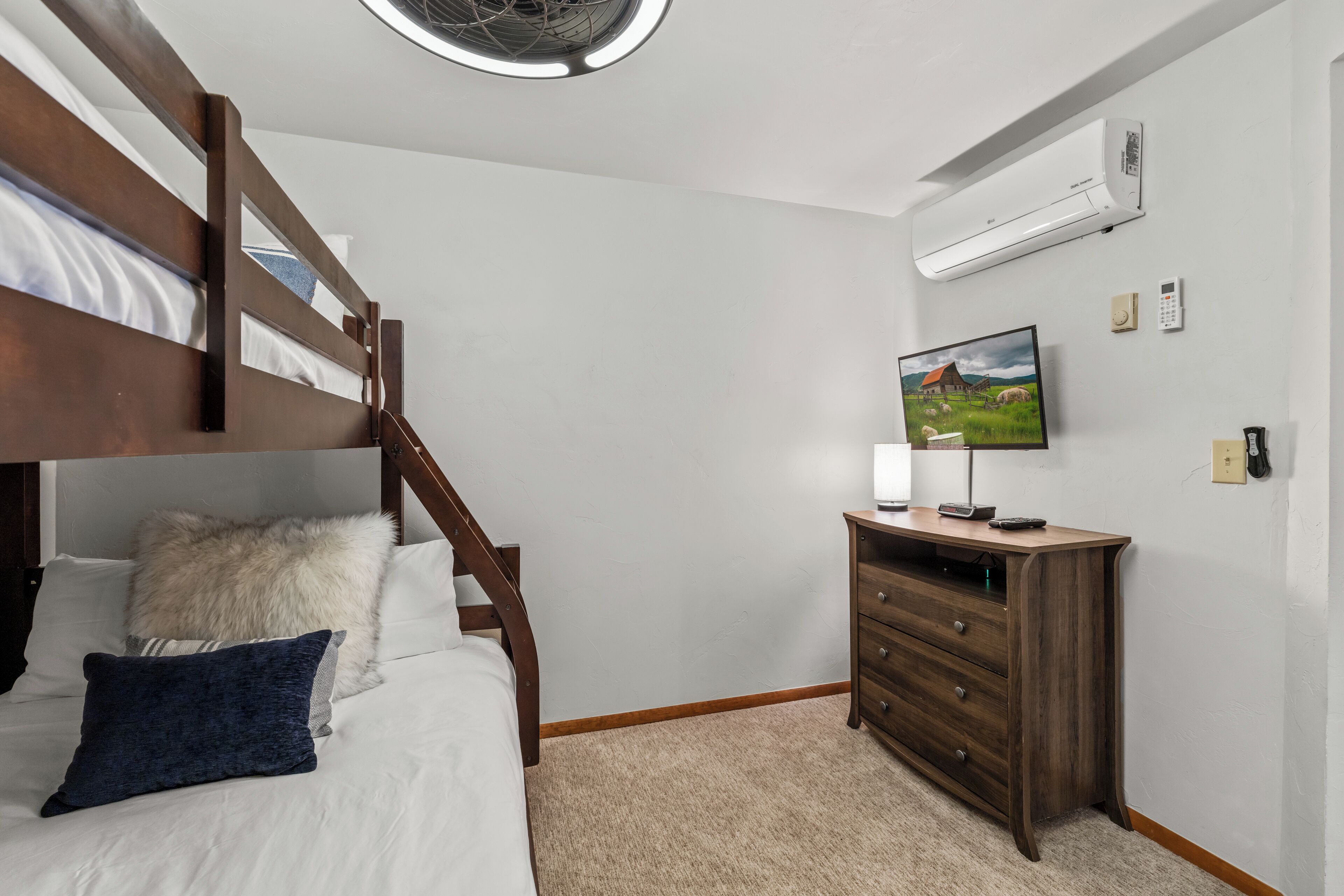 Une chambre avec un lit superposé, une commode en bois, un téléviseur à écran plat et un climatiseur fixé au mur.