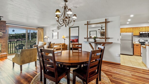 A spacious dining area with a wooden table and chairs, a chandelier, and a view of the outdoors through large windows. - Bear Claw 208 - Mountain Condo With Ski-In/Ski-Out Access! (Steamboat Springs)