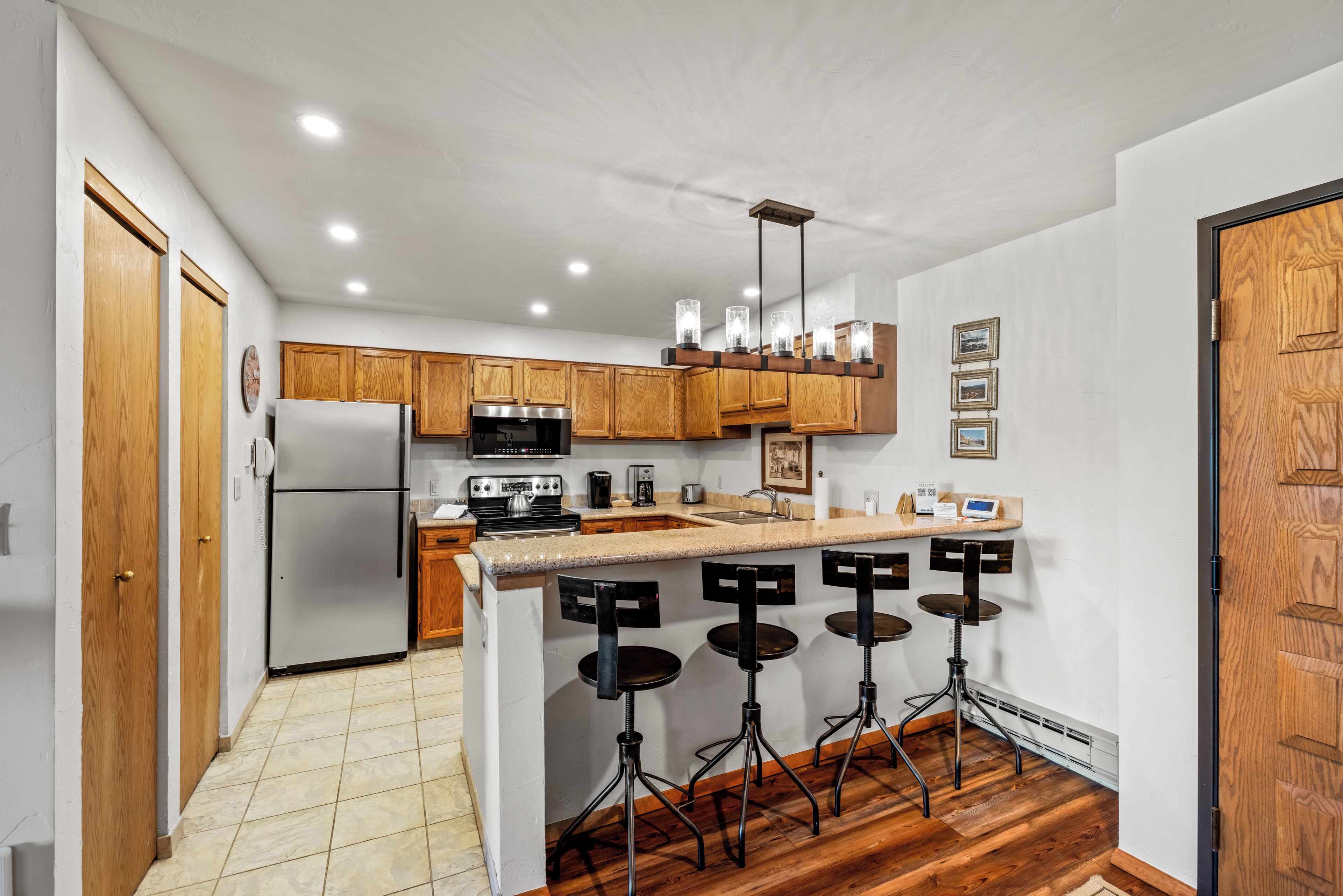 A modern kitchen with wooden cabinets, a stainless steel refrigerator, a microwave, and a coffee maker. There are three stools at the bar counter.