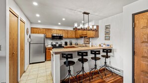 A modern kitchen with wooden cabinets, a stainless steel refrigerator, a microwave, and a coffee maker. There are three stools at the bar counter. - Bear Claw 208 - Mountain Condo With Ski-In/Ski-Out Access! (Steamboat Springs)