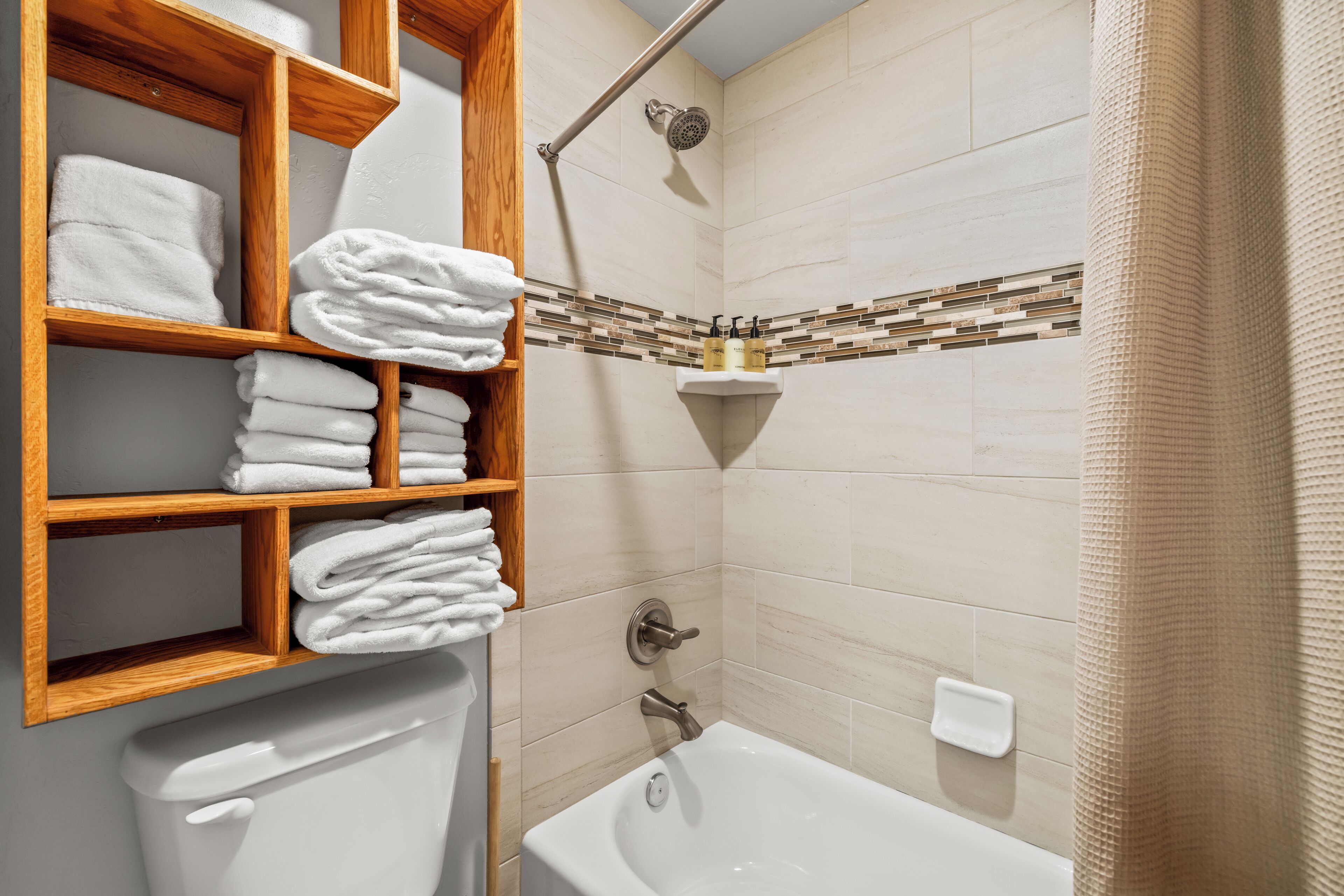 Une salle de bain comprenant des toilettes, une baignoire, une douche et une étagère en bois où sont soigneusement pliées des serviettes.