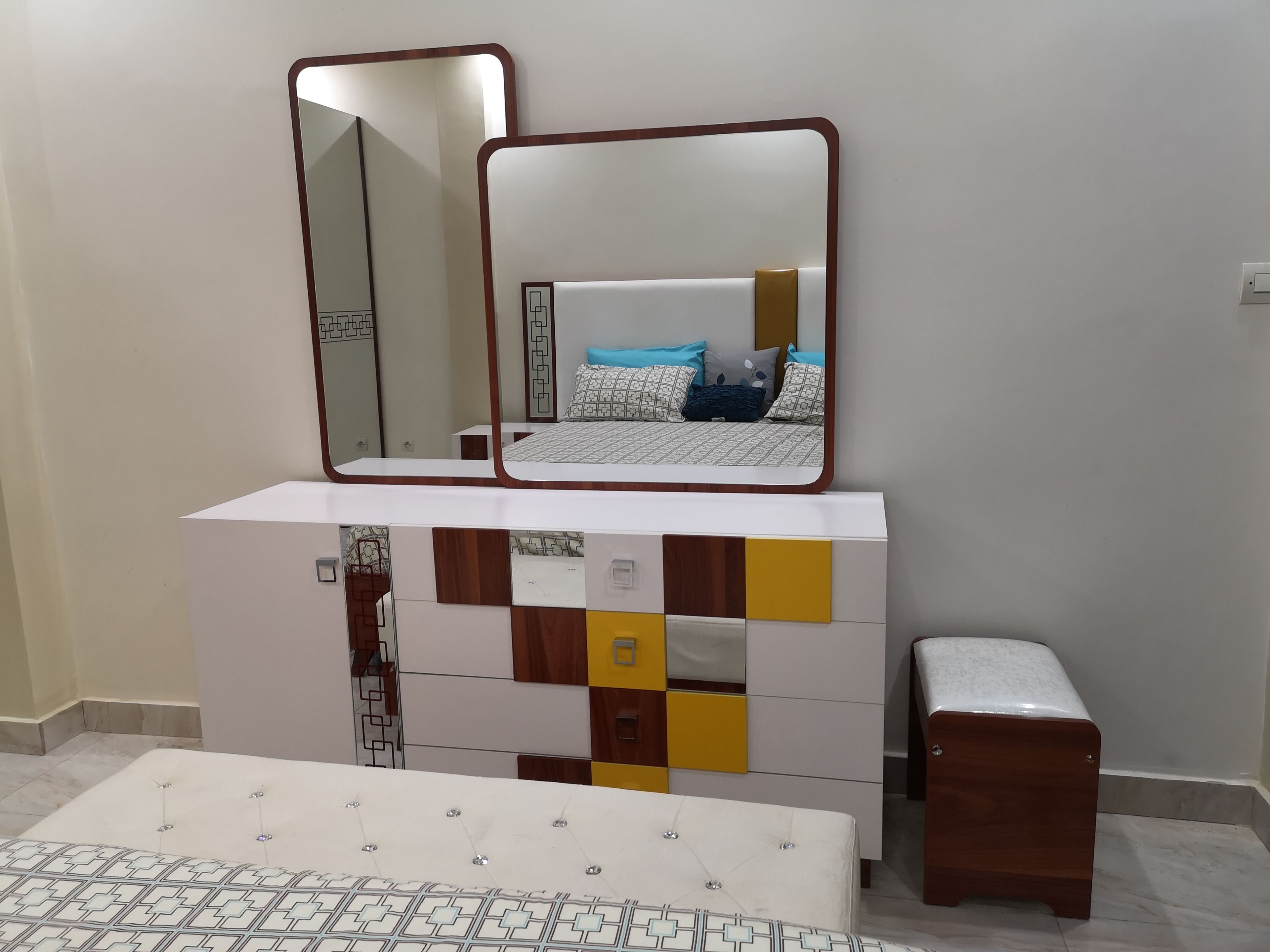 A bedroom with a bed, a bedside table, a dresser with mirrors, and a small stool.